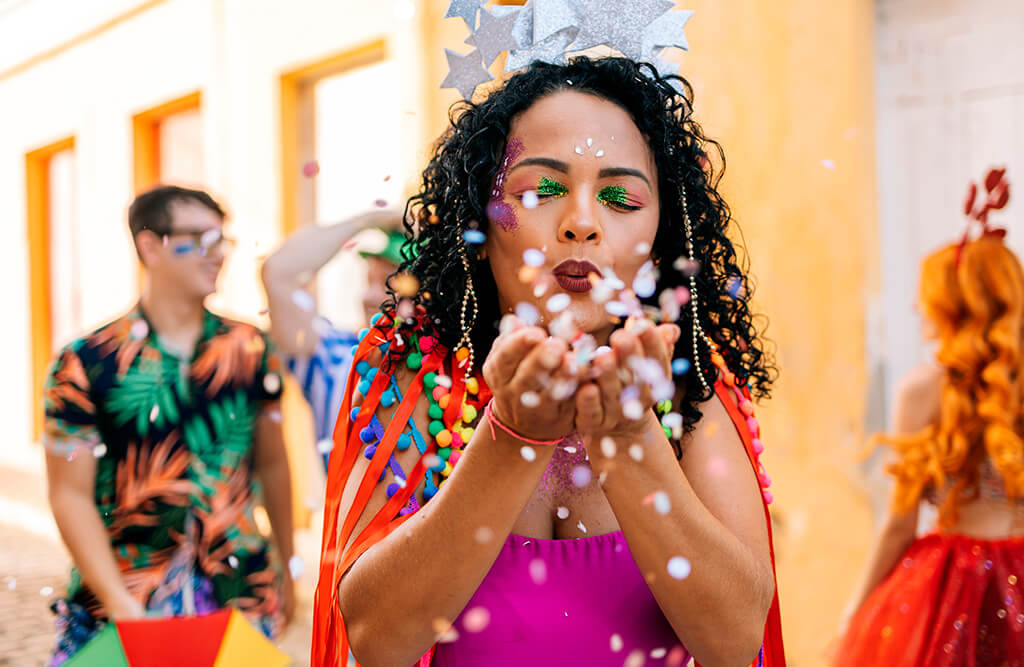 Mulher fantasiada soprando confetes durante a festa de Carnaval de 2025, cercada por foliões em um ambiente festivo e colorido.