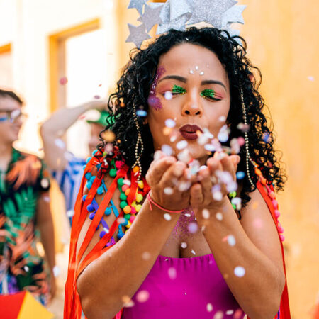 dicas-carnaval-2025-festa-folioes Mulher fantasiada soprando confetes durante a festa de Carnaval de 2025, cercada por foliões em um ambiente festivo e colorido.